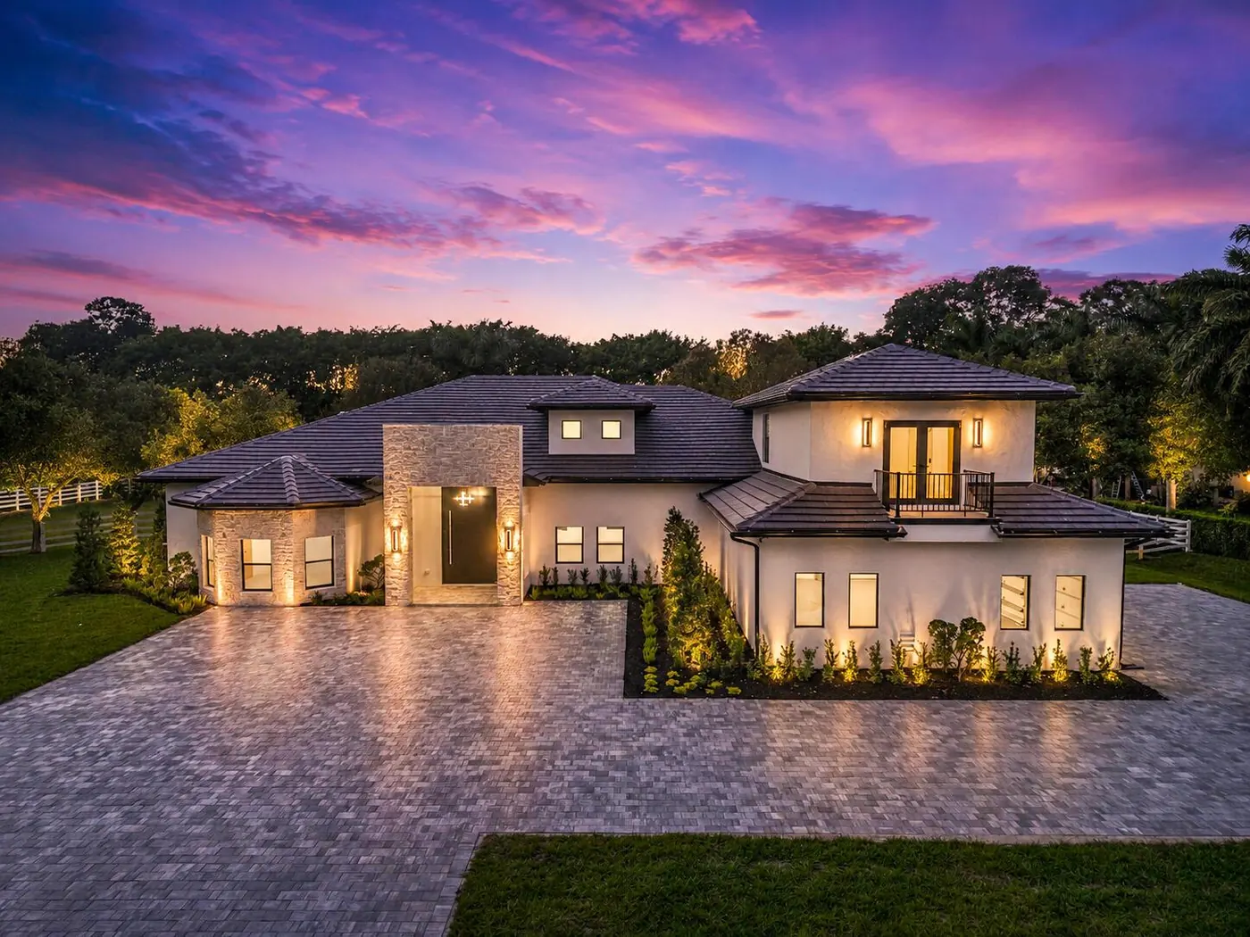 Modern two-story estate lit at dusk with a paver driveway